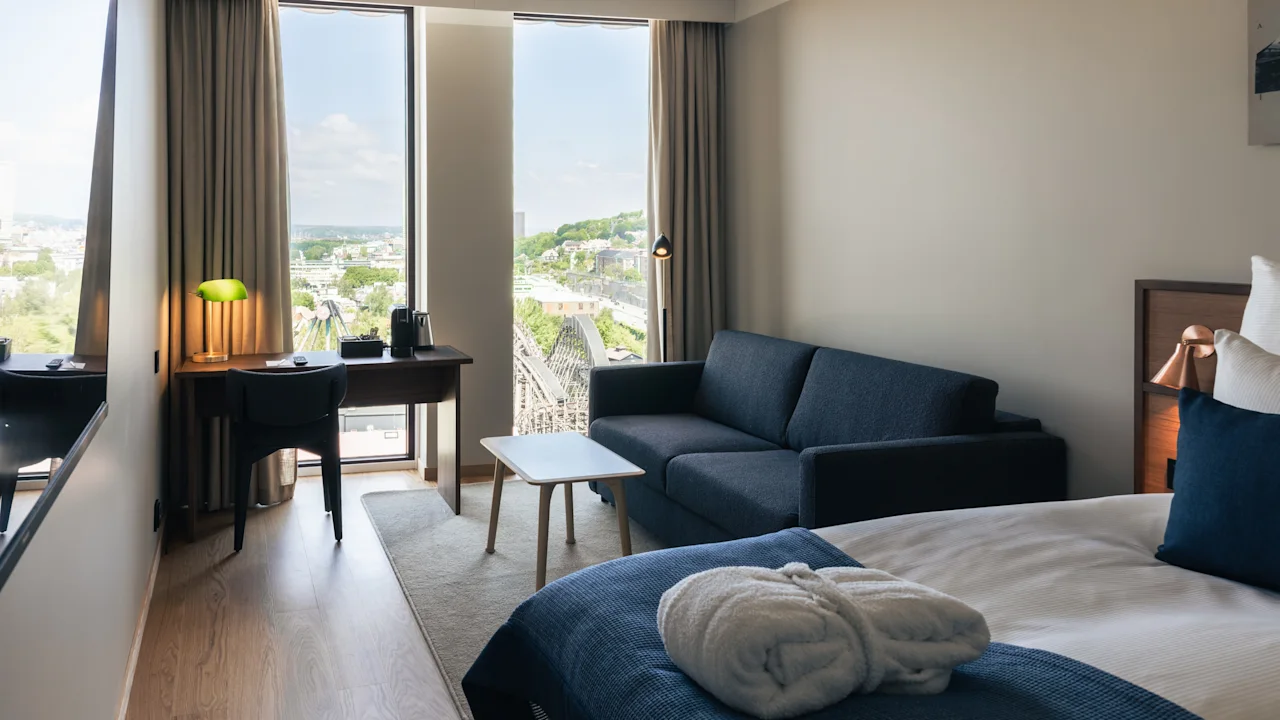 Ett ljust och inbjudande hotellrum med en mysig säng, en soffa, ett skrivbord och stora fönster med stadutsikt.