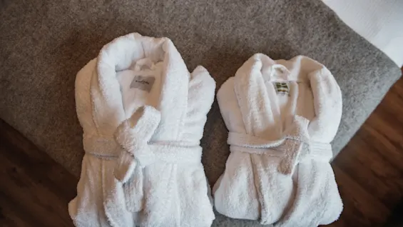Två fluffiga vita badrockar prydligt vikta på en mjuk grå filt, inbjudande till komfort och avkoppling.