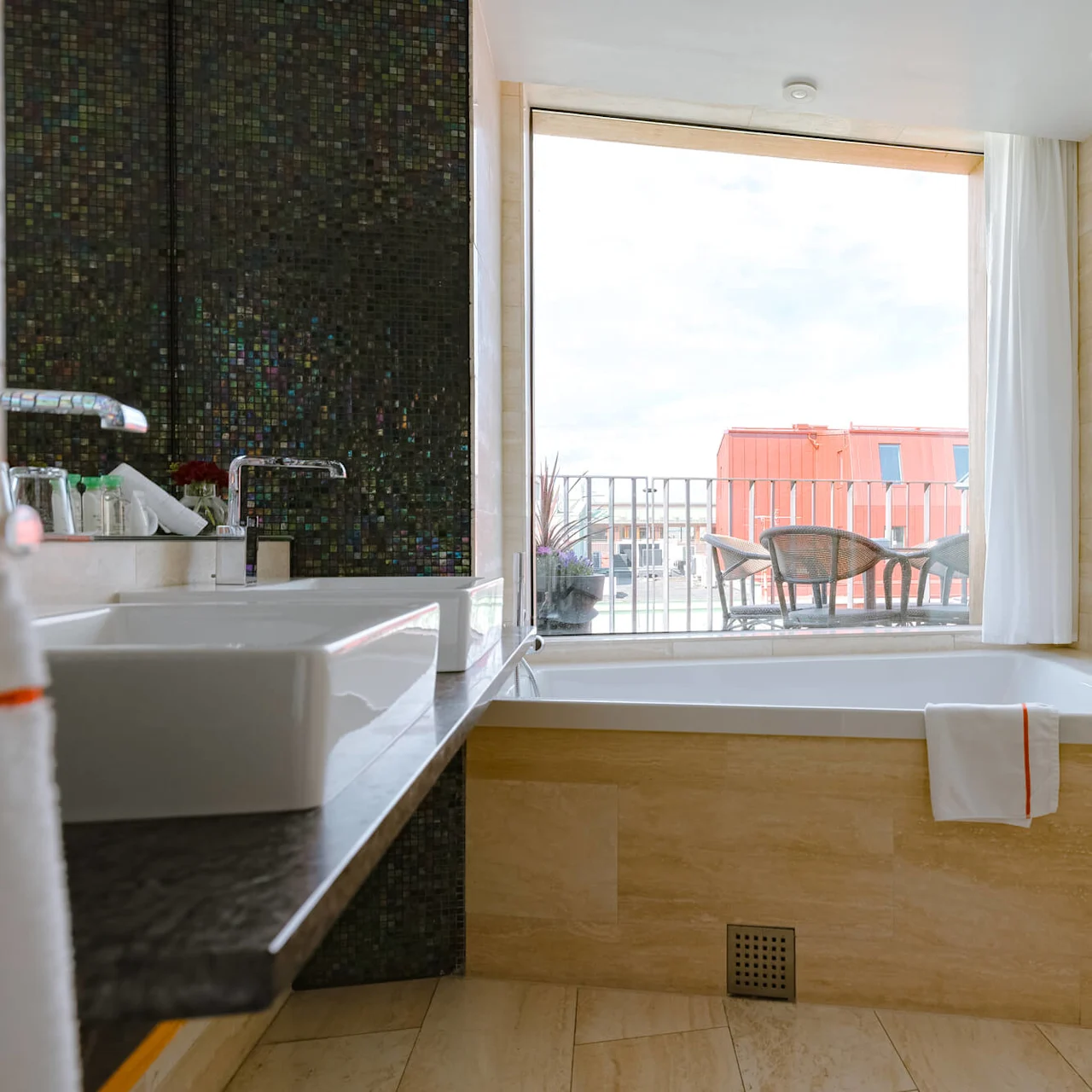 A modern bathroom with a large bathtub below a window, surrounded by beige tiles. Outside, wicker chairs and a table sit on a balcony, overlooking a red building.