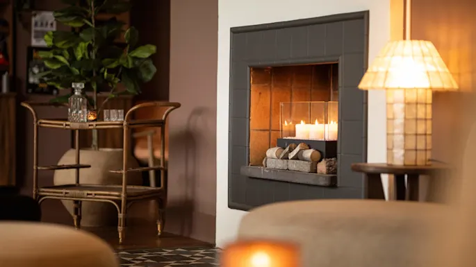 En peis fylt med tente lys kaster en varm glød i et koselig rom med en kurvvogn med glass og en høy potteplante, som forsterker den intime atmosfæren.