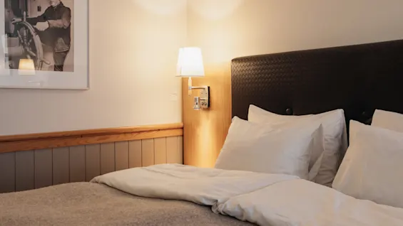 A cozy hotel room with a made bed, white linens, and a dark headboard. A framed picture hangs on the wall.