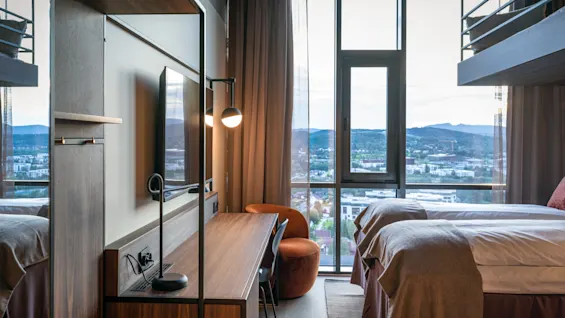 Et moderne hotelværelse med to senge, et skrivebord og et stort vindue med naturskøn udsigt over byen og bjergene.