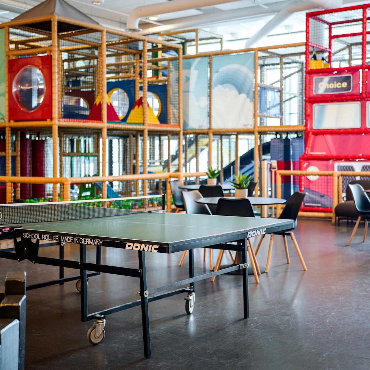 Ping pong table on wheels in recreational room; surrounded by chairs, tables, play structures, and colorful wall designs. Text: "DONIC," "SCHOOL ROLLER MADE IN GERMANY." 