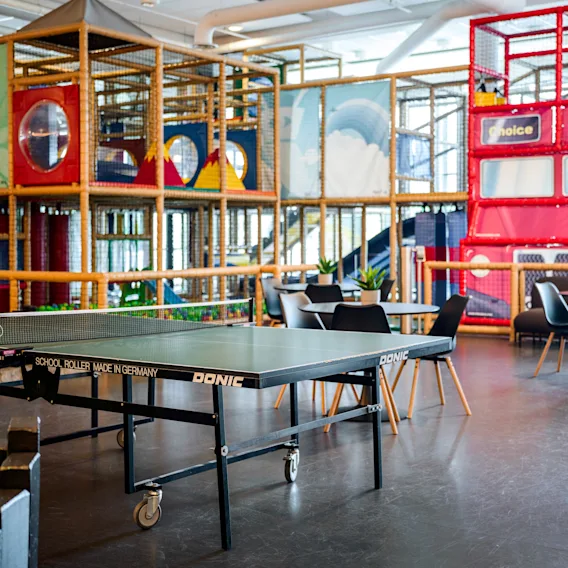 Bordtennisbord på hjul i rekreasjonsrom; omringet av stoler, bord, lekestrukturer og fargerike veggdesigner. Tekst: "DONIC," "SCHOOL ROLLER MADE IN GERMANY."