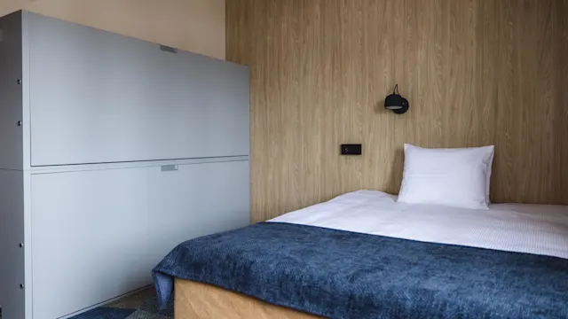 Säng står prydligt i ett enkelt rum med väggpaneler i trä och ett blått täcke. En modern grå garderob står intill och förstärker den minimalistiska inredningen.
