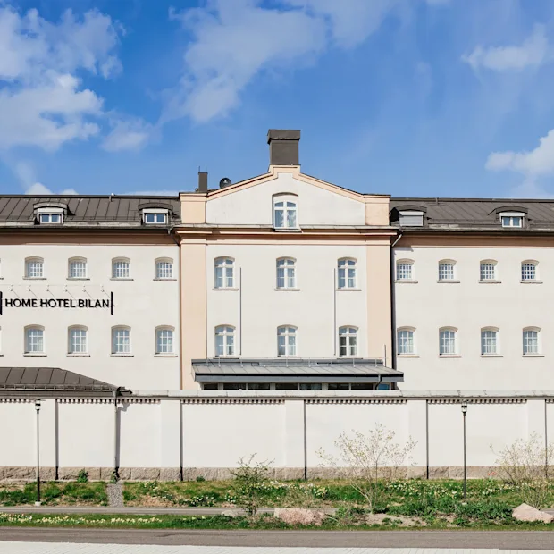 A large, light-colored building with many windows and a dark roof under a blue sky. A white wall stands in front of it. Text on building: HOME HOTEL BILAN.