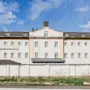 A large, light-colored building with many windows and a dark roof under a blue sky. A white wall stands in front of it. Text on building: HOME HOTEL BILAN.