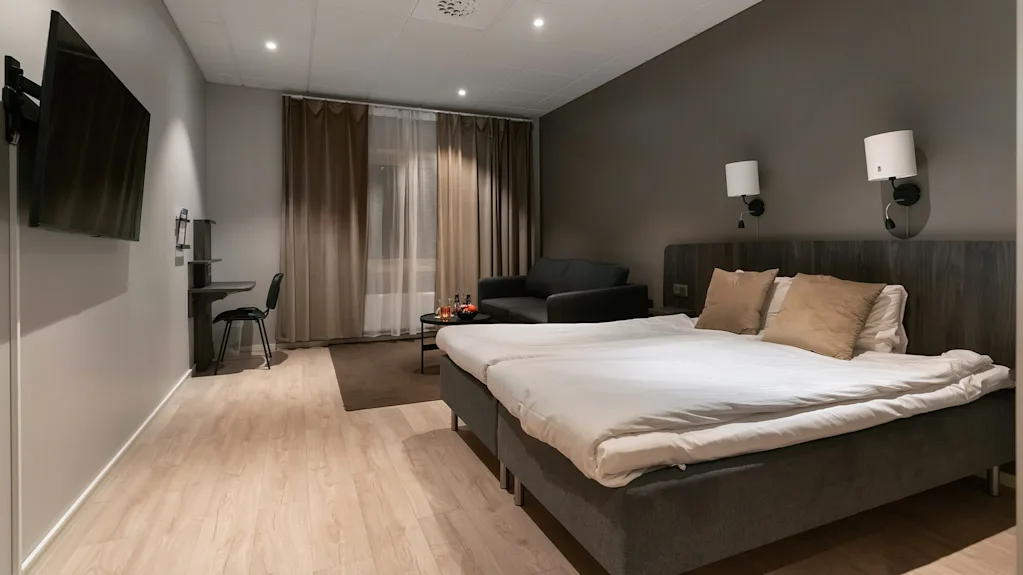 En prydligt bäddad säng med kuddar står bredvid vägglampor i ett modernt hotellrum. Till vänster finns ett skrivbord med stol och TV, med en soffa och gardiner bakom.