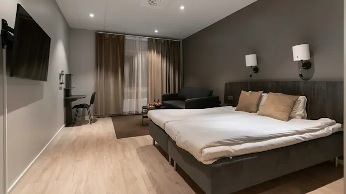 En pent oppredd seng med puter står ved siden av vegglamper i et moderne hotellrom. Et skrivebord med stol og TV er til venstre, med en sofa og gardiner bak.