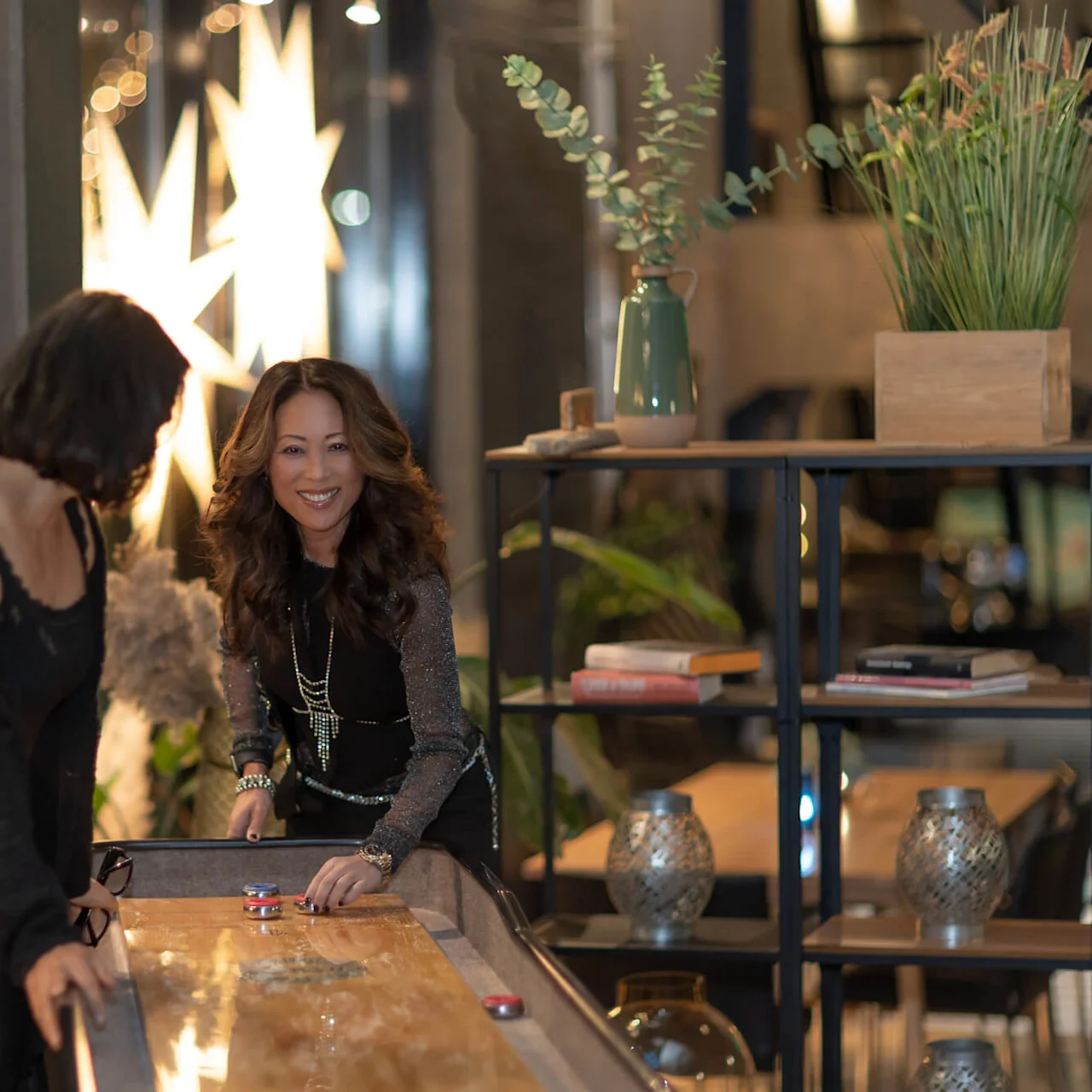 Two women play shuffleboard, one smiling while pushing a puck. The room is decorated with plants and books, softly lit by star-shaped lights.