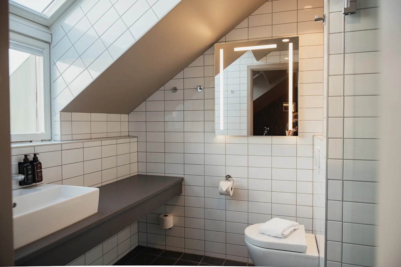 Et lyst, moderne badeværelse med hvide fliser, skråloft og et oplyst spejl, der skaber et rent og funktionelt rum.