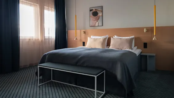 A neatly made double bed with gray bedding sits in a modern bedroom. Two pendant lamps hang beside it. A bench is at the foot, and curtains cover a window.