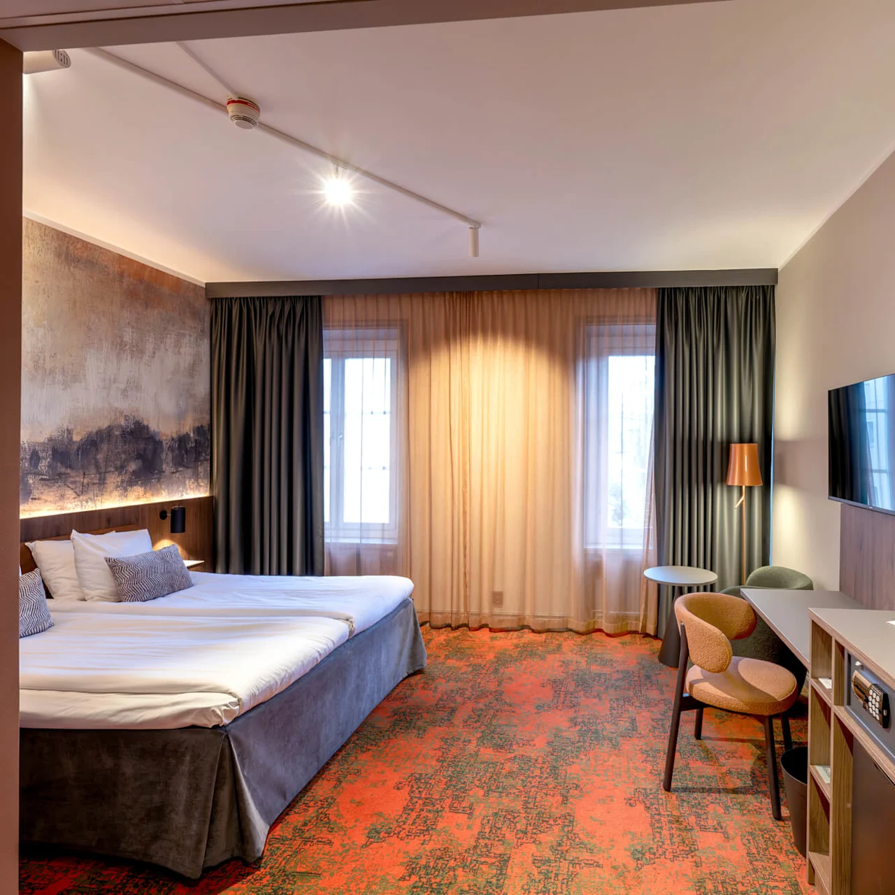A well-furnished hotel room features a large bed, wall art, and a desk area with chairs. It is brightly lit with floor-to-ceiling curtains and a wall-mounted TV.
