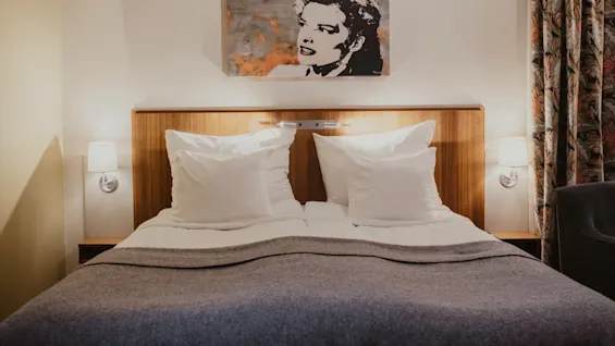 A cozy hotel room at Home Hotel Kung Oscar with a neatly made bed, white pillows, and a grey blanket. Artwork hangs above the bed.