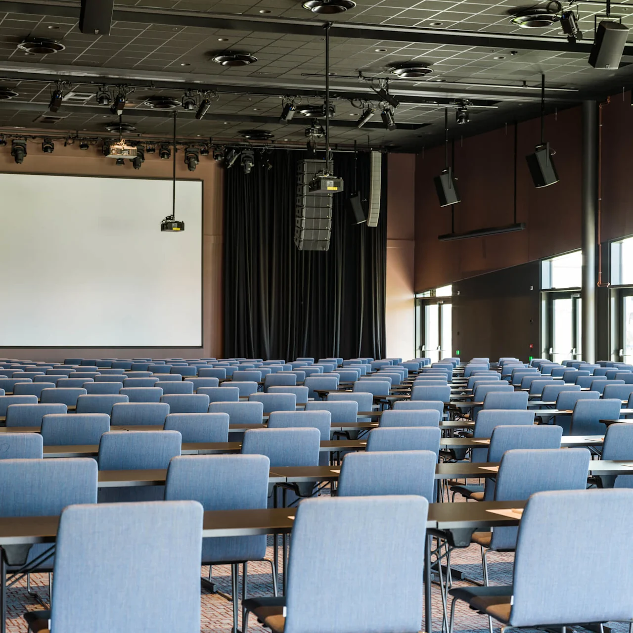 Rader med tomme blå stoler vender mot en stor tom skjerm, omgitt av svarte gardiner i et romslig konferanselokale.