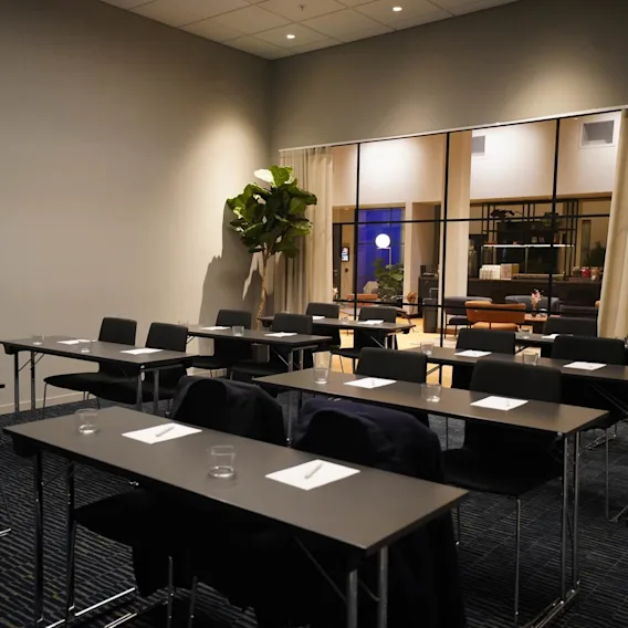 Rows of empty conference tables with chairs, paper, and glasses, in a room with a potted plant and adjoining area visible through large windows, under soft lighting.
