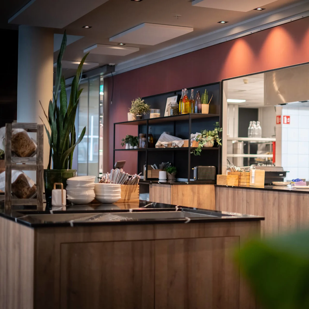 A modern restaurant kitchen with wooden countertops features neatly arranged dishes and utensils. Potted plants and industrial lights add warmth, while the open design offers a view of the cooking area.