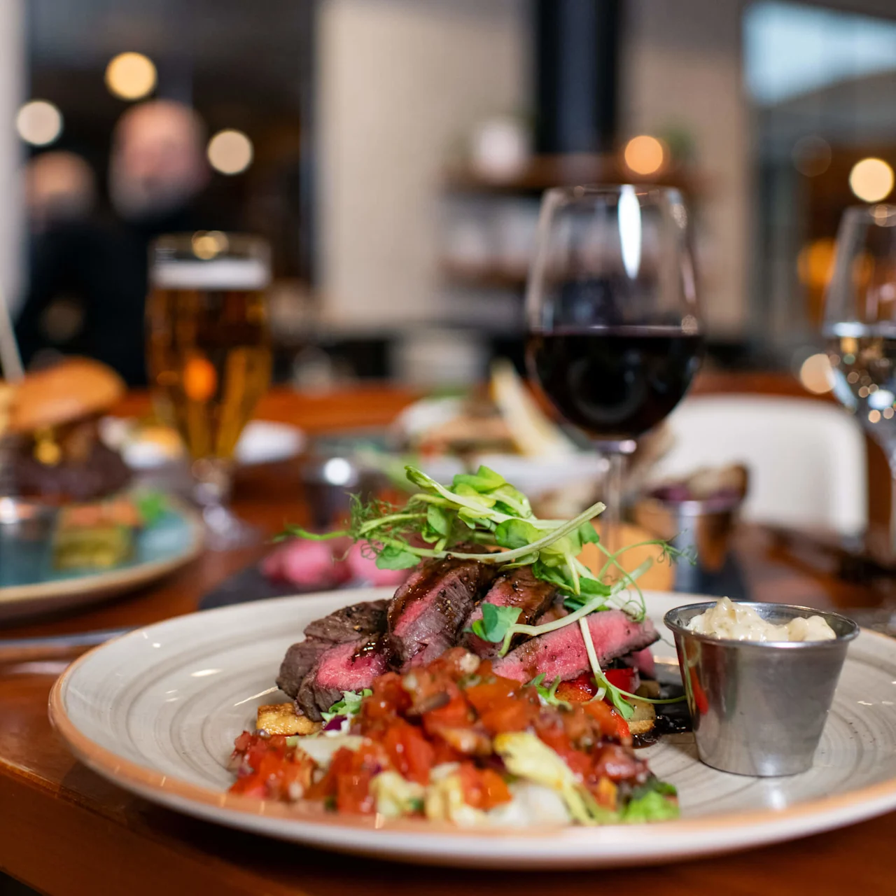 A plate of sliced steak topped with greens is served with roasted vegetables and a sauce cup. Two glasses, one of red wine and one of beer, sit nearby in a restaurant setting.