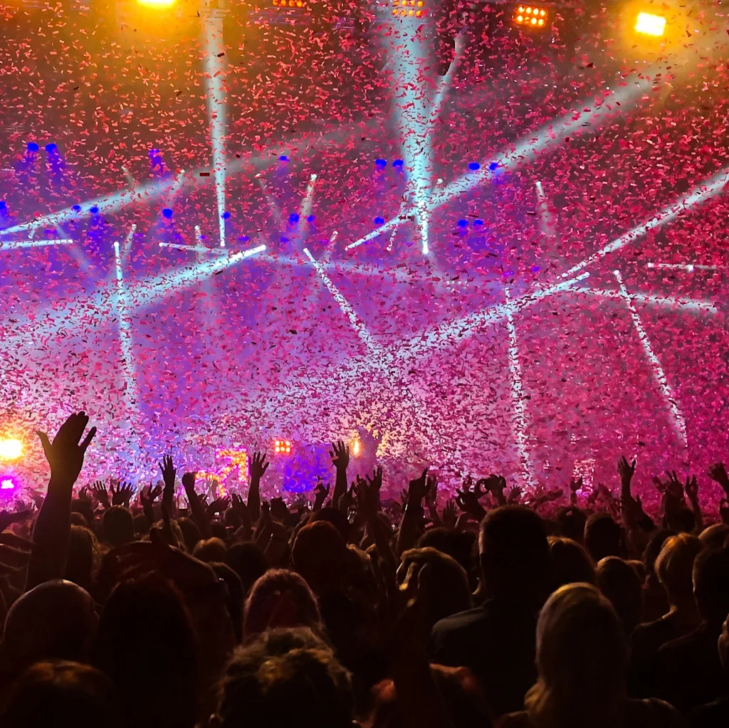 En livlig konsertscen med en folkmassa, händer uppsträckta, under starka scenljus och ett regn av färgglad konfetti.