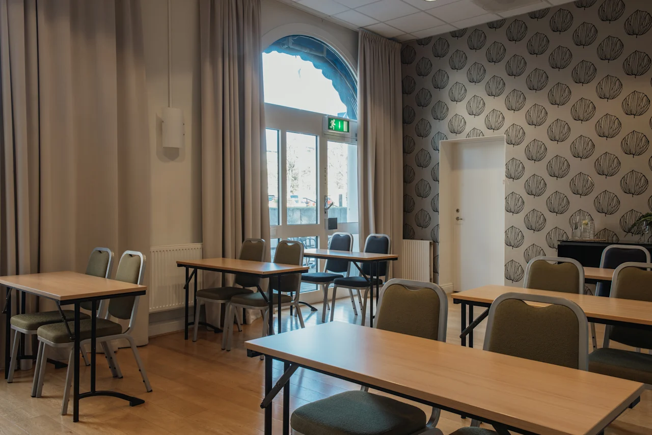 Ett ljust mötesrum med flera bord och stolar, ett stort välvt fönster och mönstrad tapet. EXIT-skylt synlig.