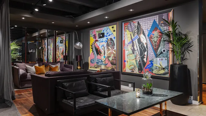 Modern living room with abstract paintings on the wall, a large dark sofa, and a coffee table. Environment features wooden flooring and dim overhead lighting, creating a cozy atmosphere.