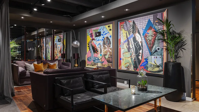 Modern living room with abstract paintings on the wall, a large dark sofa, and a coffee table. Environment features wooden flooring and dim overhead lighting, creating a cozy atmosphere.