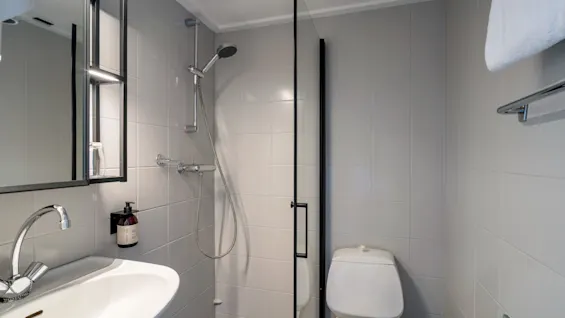 A small, modern bathroom features a wall-mounted sink with a mirror above, a shower enclosed with glass, and a toilet beside it. The walls are tiled light gray.