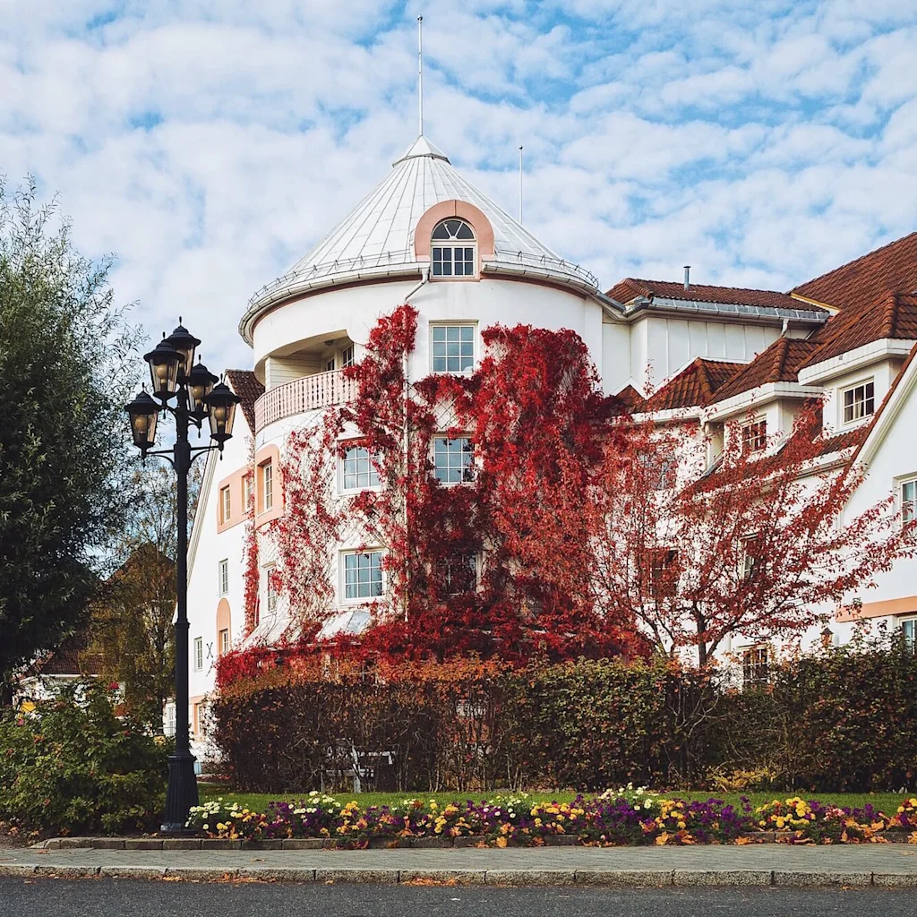 En vit byggnad med ett runt torn är prydd med röda klätterväxter. Den står bredvid en gatlykta på en blomsterkantad gata, under en delvis molnig himmel.