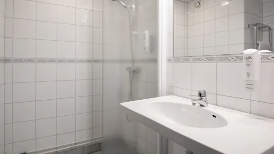 Dusch med frostat glasdörr bredvid en vit vask, i ett kaklat badrum. En tvålpump märkt "LUX Hotel Soap 450ML" är monterad på väggen.