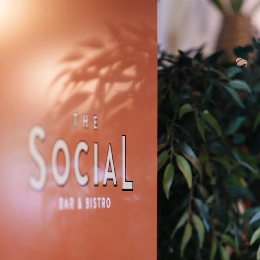 En skylt lyder "The Social Bar & Bistro" mot en orange bakgrund, med bladiga växter delvis synliga på sidan.