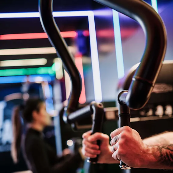 Händer som greppar gymutrustningshandtag, tränar i ett neonupplyst gym. En person syns i bakgrunden och använder en liknande maskin.