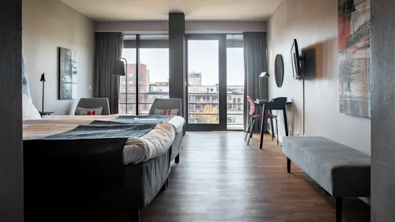 A hotel room with a neatly made bed, a desk, and a chair, overlooks a cityscape through a large window, featuring modern décor and minimalistic design.