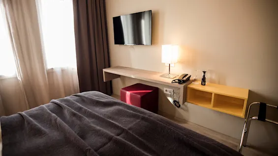 Vegghengt TV henger over et skrivebord med lampe, telefon og vinglass, ved siden av et vindu med gardiner, i et enkelt hotellrom.