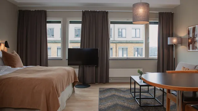 Säng täckt med en beige filt står bredvid en TV på ett stativ. Bord, stolar och en lampa placerade bredvid i ett rum med tre stora fönster och gardiner.