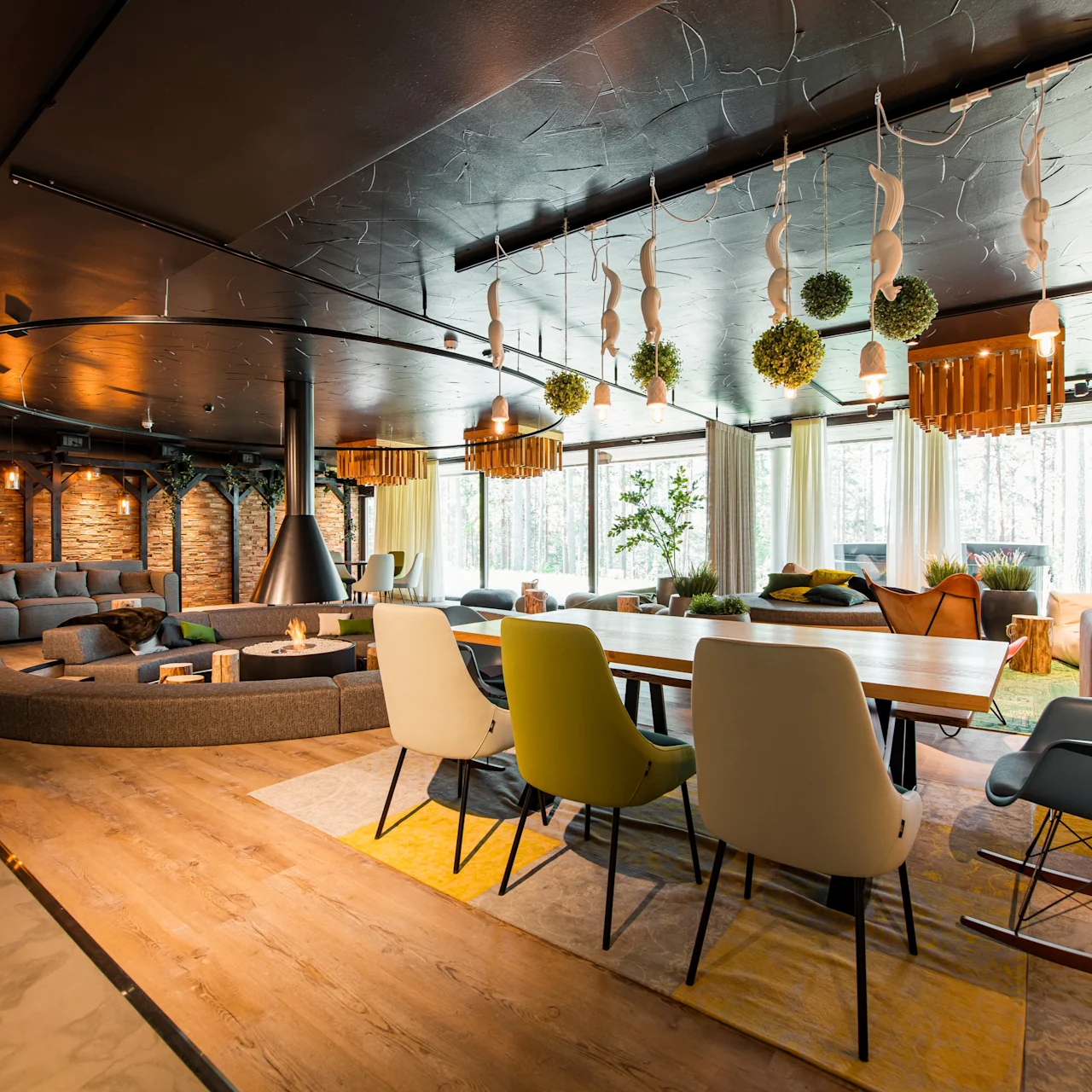 Modern living space featuring a large wooden dining table, colorful chairs, and a central fireplace. Surrounded by soft couches, large windows offer views of a forested area.