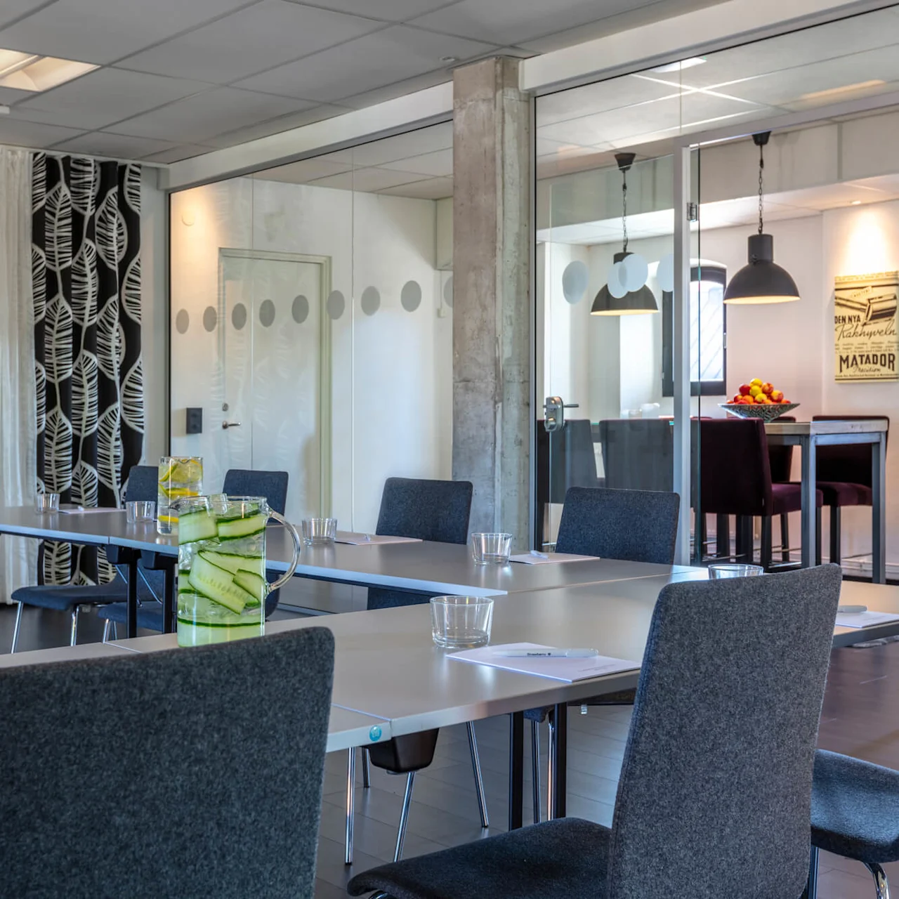 A modern meeting room with a long table, chairs, and refreshments, featuring glass walls and a poster on the back wall. 