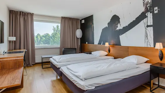 Hotellihuone, jossa on kaksi erillistä vuodetta, puinen pöytä ja suuri ikkunanäkymä vihreille puille. Seinällä sängynpäätyjen yläpuolella muraali henkilöstä koskemassa ovenkahvaa.