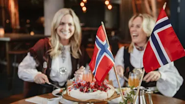 To kvinner i bunad ler og skjærer i en kake dekorert med norske flagg, i en koselig restaurantinnstilling.