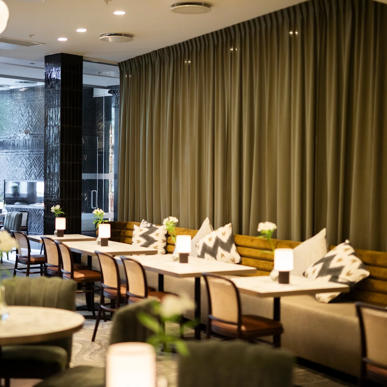 Tables and chairs arranged in a cozy, modern restaurant. Soft lighting and decorative pillows enhance intimate ambiance. Tables hold small lamps and vases with flowers, adding elegance to the space.