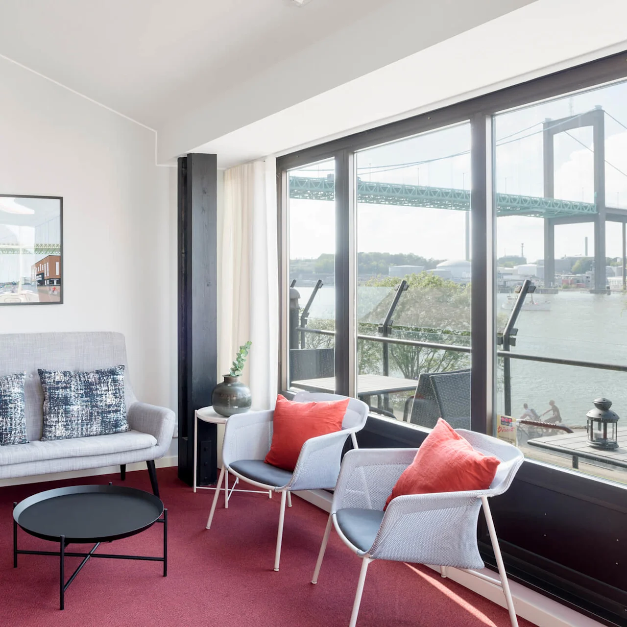 A small, modern seating area with gray chairs and a sofa surrounds a black table. Large windows reveal a bridge and waterway outside. Pendant lights hang from the ceiling.