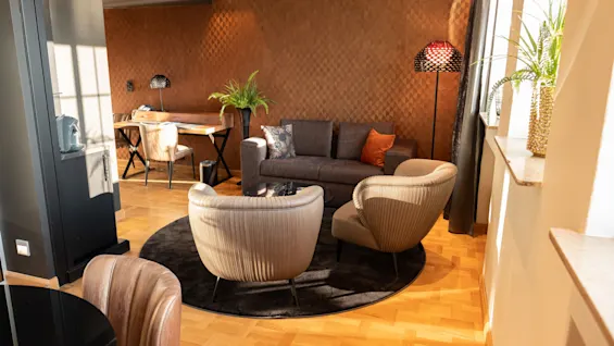 En stue har to beige lenestoler på et sirkulært teppe, vendt mot en brun sofa. Et tregulv kontrasterer med en teksturert oransje vegg. Et spisebord og planter pynter rommet.