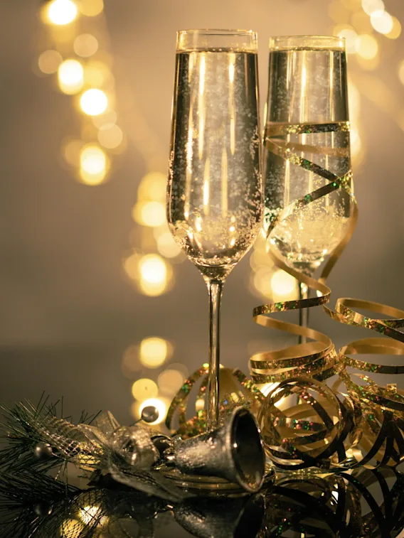 To champagneglass står sammen, omgitt av gyldne bånd og festlige dekorasjoner på en reflekterende overflate, med myke, varme lyslenker som gløder i bakgrunnen.