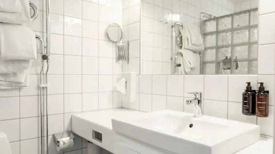 Ett ljust, vitkaklat badrum med handfat, spegel och toalettartiklar märkta TVÅL och BODY LOTION. Färska handdukar syns.