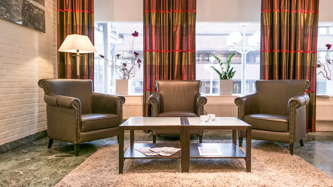 Three brown leather armchairs surround a coffee table holding magazines in a cozy lounge. A lamp stands beside, with large windows and vibrant curtains providing natural light.