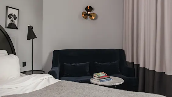 En marinblå soffa står mot en grå vägg under en modern vägglampa. Ett litet runt bord med böcker står i närheten. Gardiner täcker delvis väggen bredvid en säng.