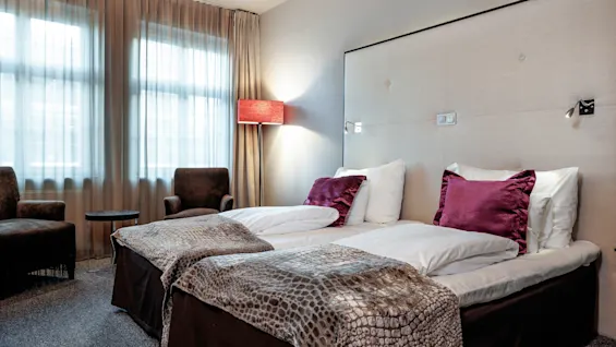 Et hotellrom har to pent oppredde enkeltsenger med mønstrede tepper og myke lenestoler. Myk belysning og store vinduer med gardiner skaper en koselig, innbydende atmosfære.
