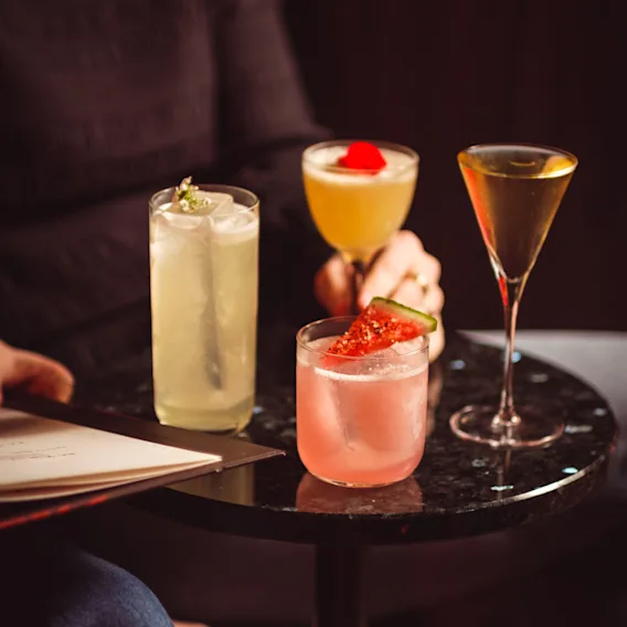 People enjoying a variety of colorful cocktails at a bar or lounge, with one person holding a drink and a menu nearby.