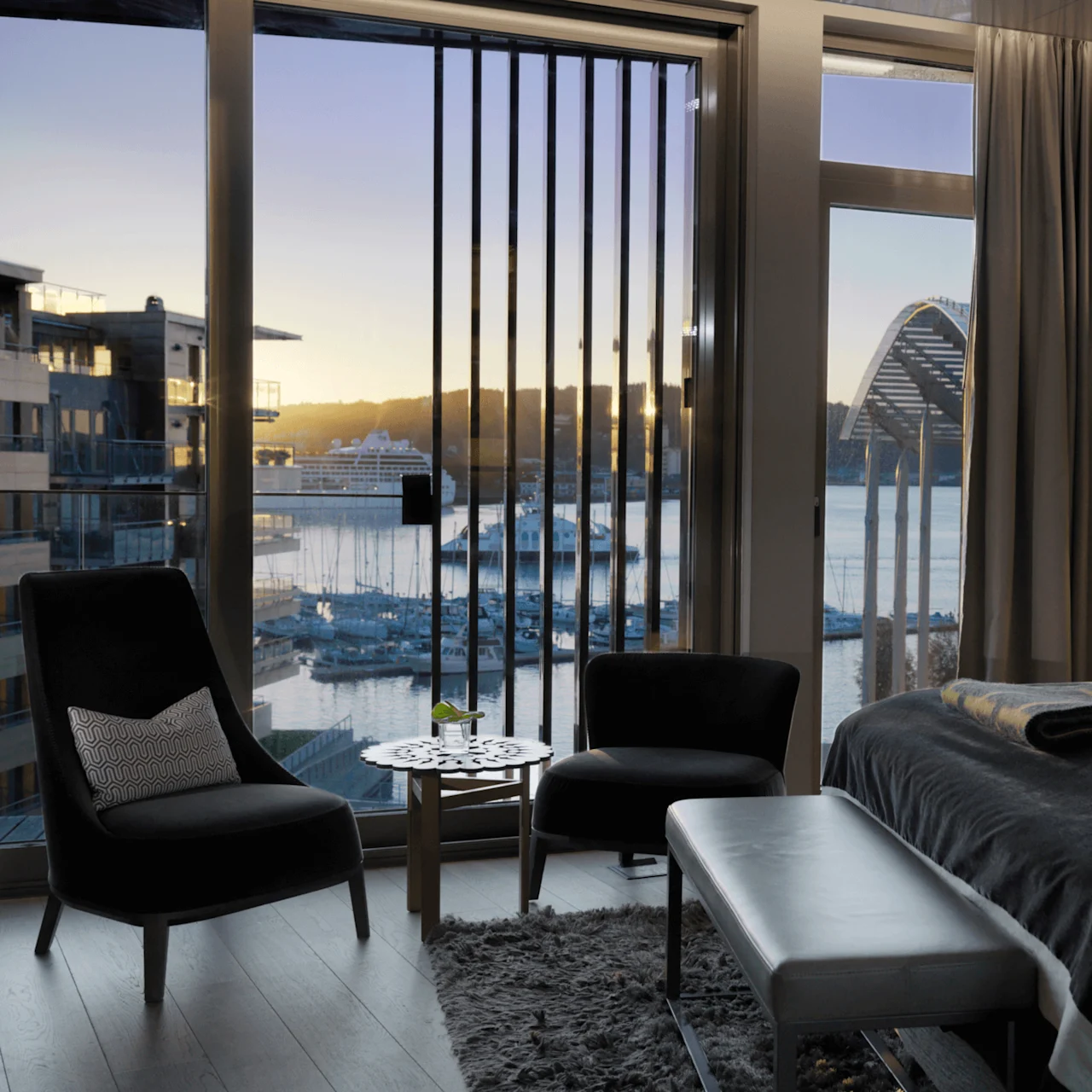 Et luksuriøst hotellrom har en seng og sittegruppe med utsikt over en marina med yachter i skumringen gjennom store vinduer. Innredningen inkluderer nøytrale toner og myk belysning.