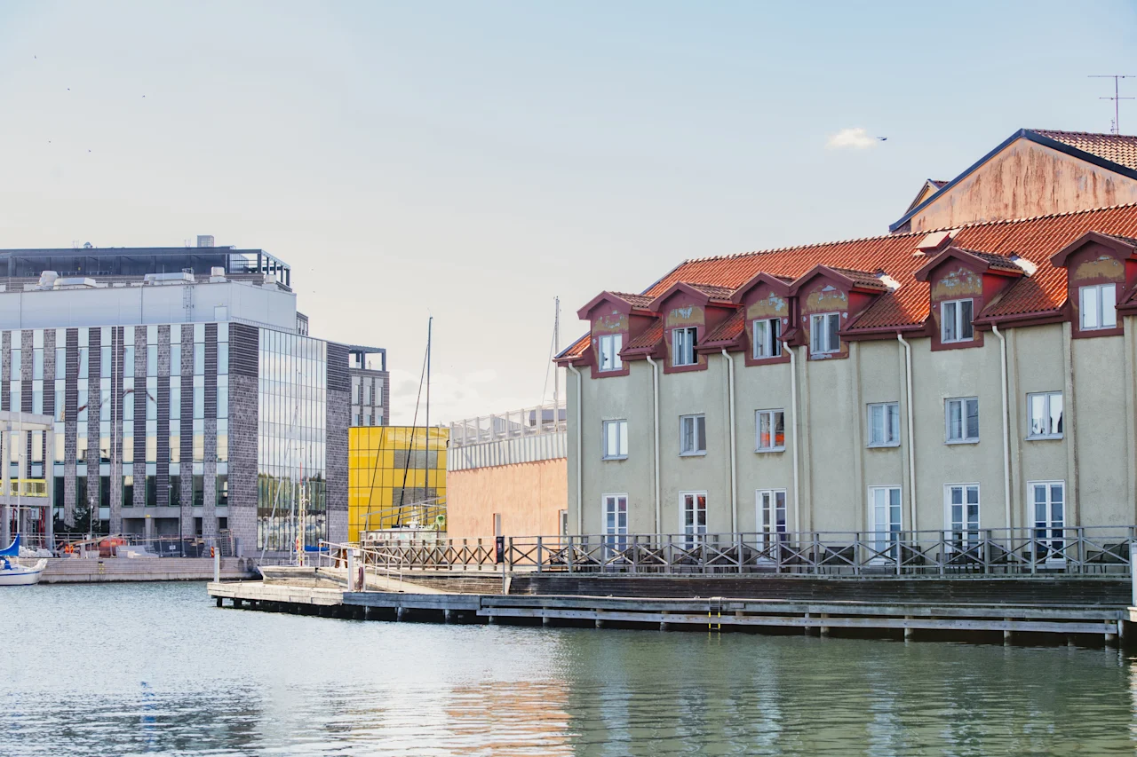 En havnefrontscene med en moderne bygning og en traditionel bygning med rødt tag, begge med udsigt over et roligt vandområde.