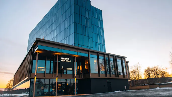 En moderne glassbygning med blå vinduer står i skumringen; et opplyst skilt lyder "HERNÖ GIN Hotell," omgitt av snødekket mark og fjerne trær under en blek himmel.